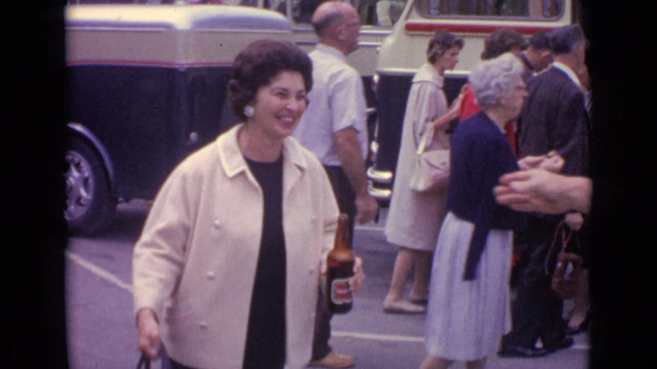 Woman in a cream jacket smiles while holding a beer bottle on a street near buses.