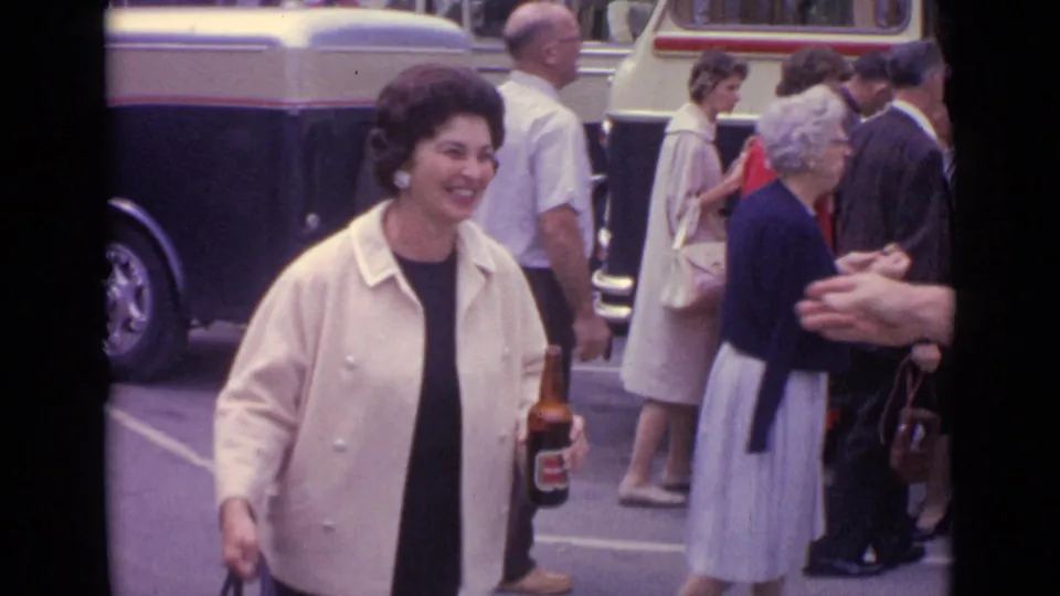 In 1962 Germany footage, a smiling woman in a cream jacket holds a beer bottle as pedestrians and buses fill the street behind her.