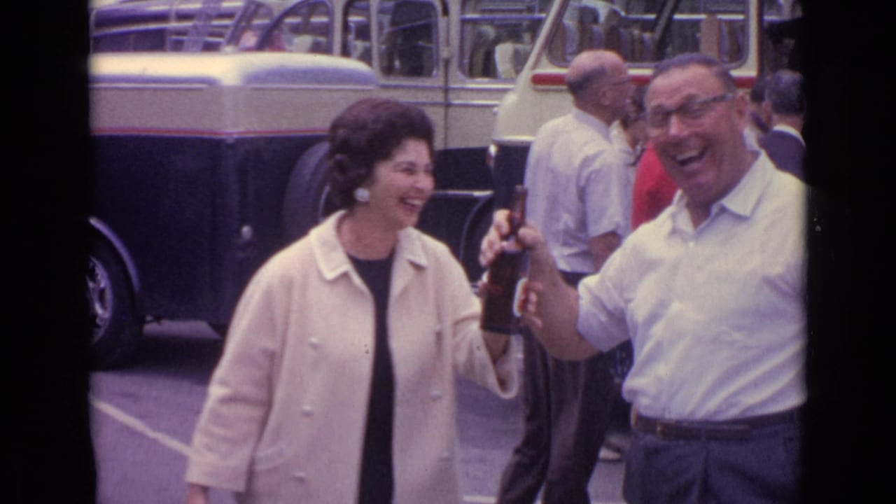 In the Germany street scene, the woman laughs while a man in a white shirt raises a beer bottle, with a bus and pedestrians behind them.