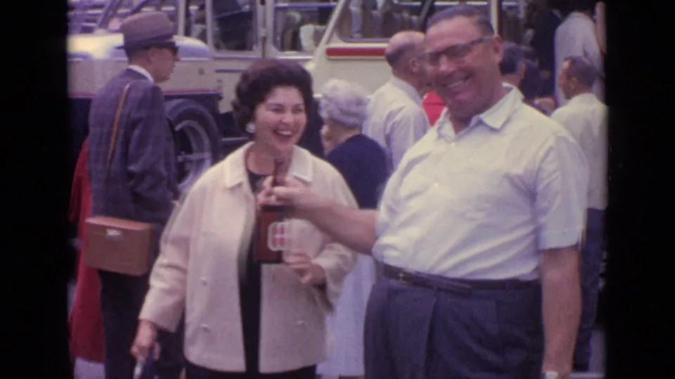 The man in a white shirt holds out the beer bottle as the woman smiles, with older pedestrians and a bus visible in the 1962 Germany street footage.