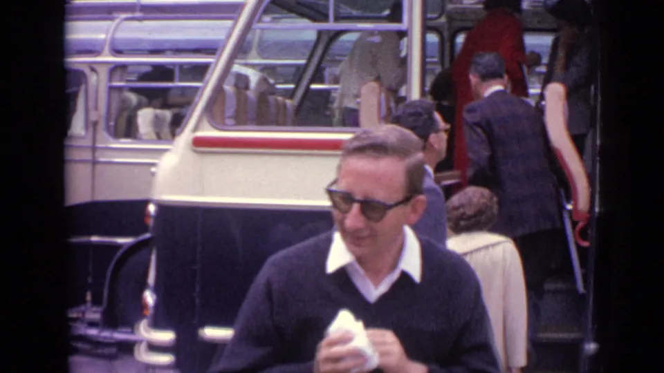 1962 Germany home-movie frame showing passengers boarding a cream and black coach with a man in sunglasses in front.