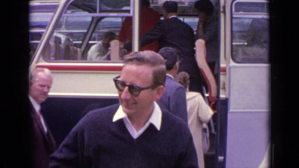 1962 Germany travel frame with a smiling man in sunglasses before a cream and black coach and passengers boarding behind him.