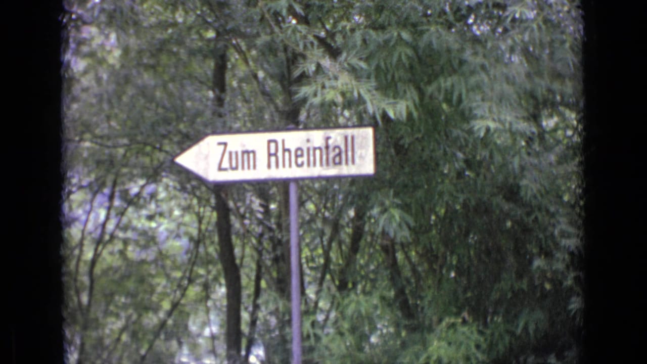 1962 8mm travel frame showing a white directional sign reading Zum Rheinfall against leafy trees.