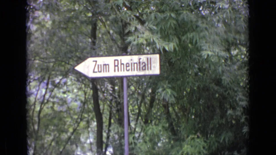1962 8mm travel frame showing a white directional sign reading Zum Rheinfall against leafy trees.