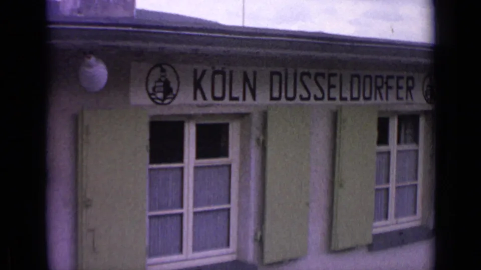 1962 8mm frame of a building exterior with KOLN DUSSELDORFER signage, windows, and green shutters.