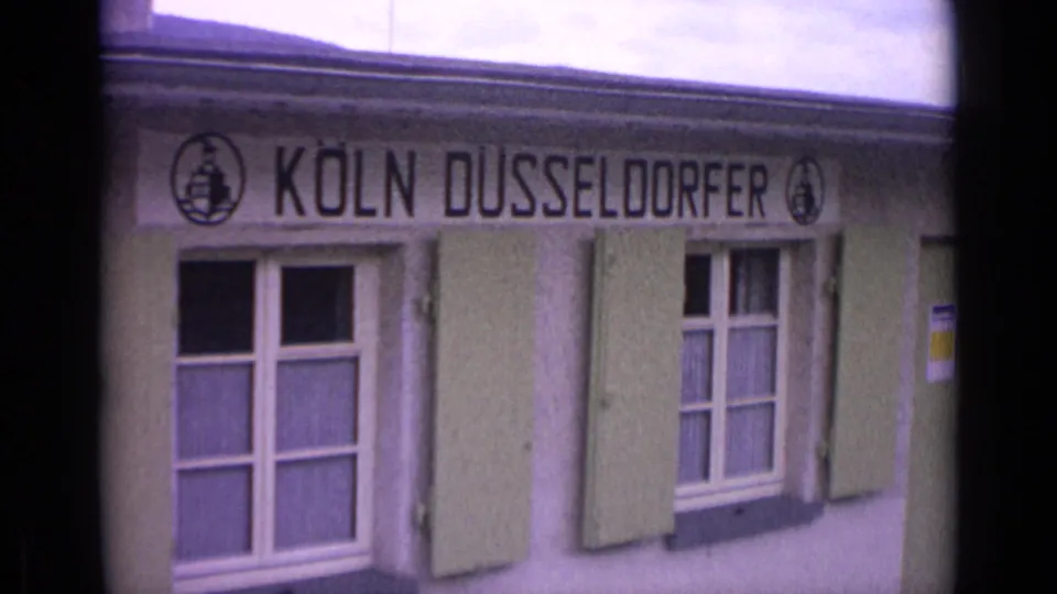 1962 8mm frame showing KOLN DUSSELDORFER facade lettering over windows with pale green shutters.