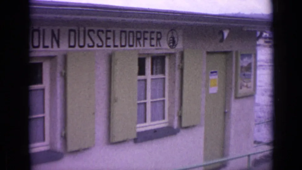 1962 Germany frame of the Köln Düsseldorfer riverside facade, with green shutters, a doorway, and railing near the edge of view.