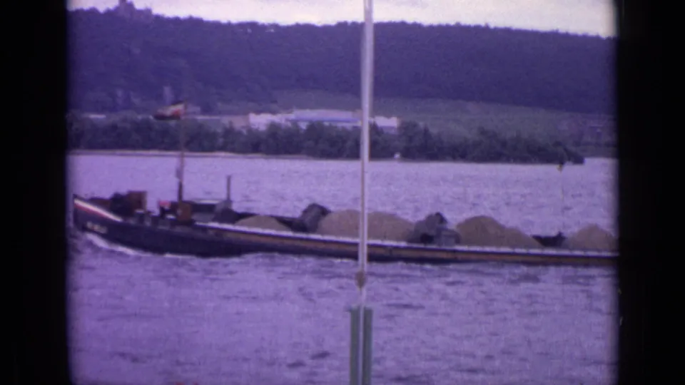 1962 Germany 8mm view of a long heavy-laden barge on the river, seen past a vertical pole with a wooded bank in the distance.