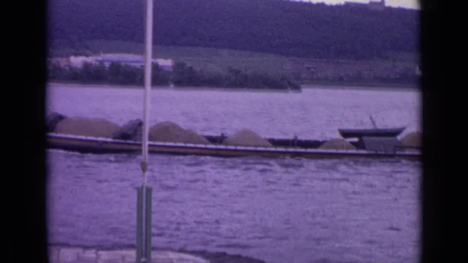 1962 Germany 8mm frame showing the loaded barge continuing along the river, with shoreline trees and fields in the background.
