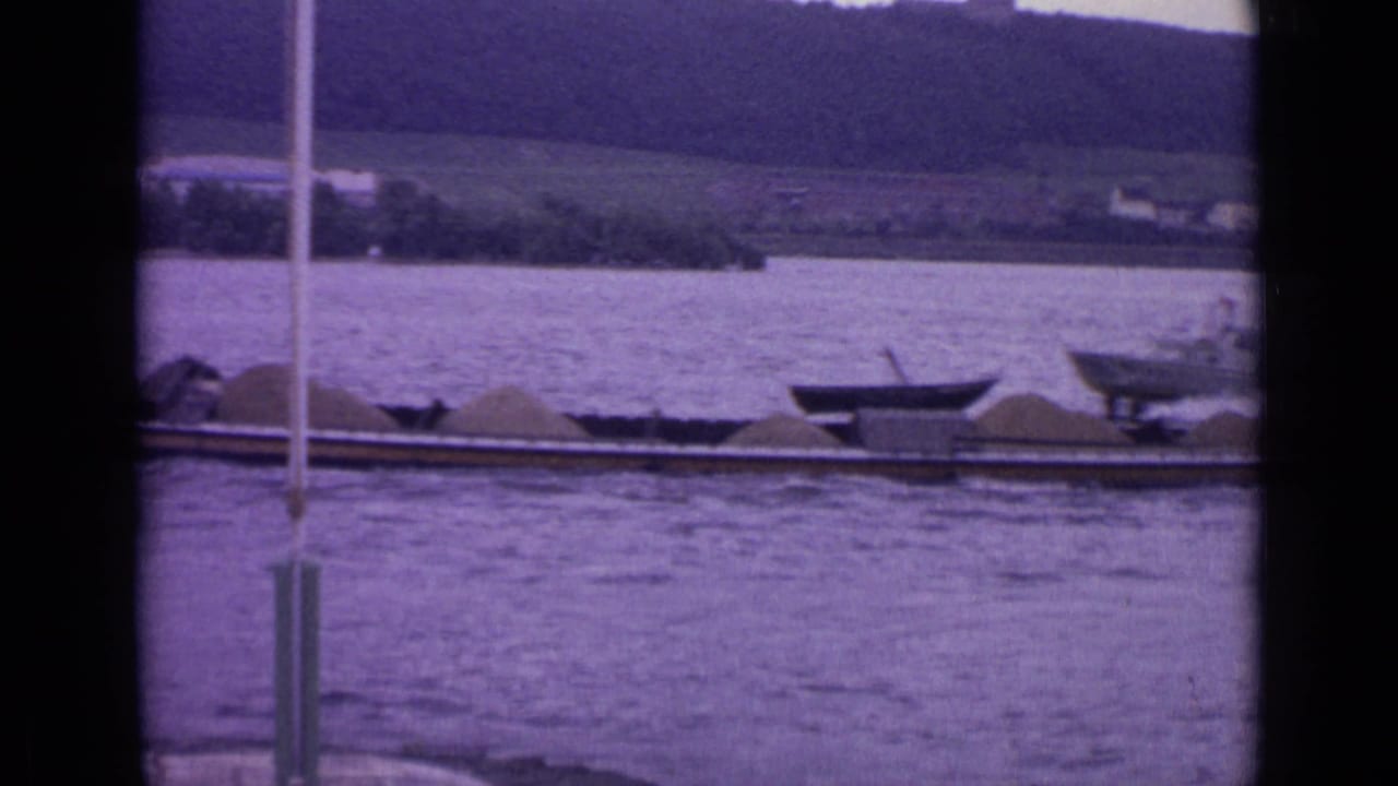 Barge loaded with pale material on a river below a wooded hillside.