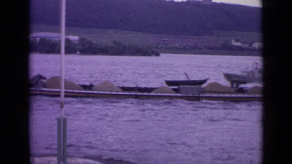 1962 Germany 8mm frame showing a low river barge with pale cargo, small boats, foreground window bars, and a wooded hillside beyond.