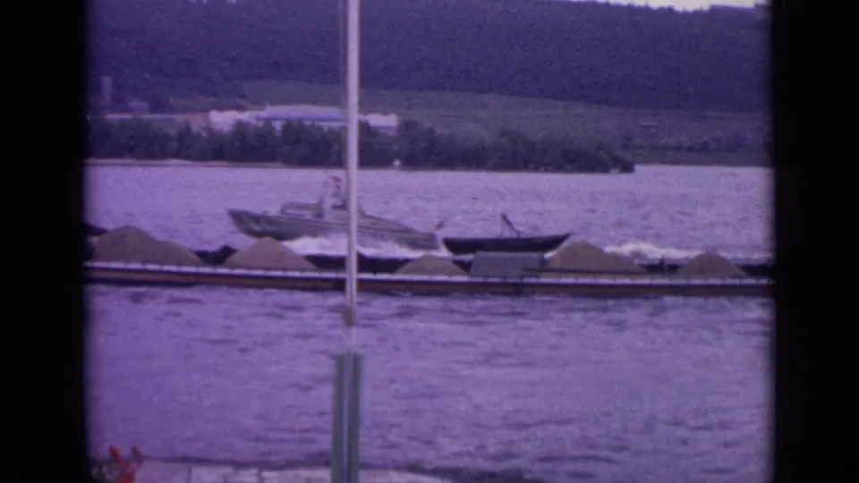 1962 Germany 8mm frame showing a motorboat wake crossing near a loaded barge, with a riverbank and wooded hillside in the background.