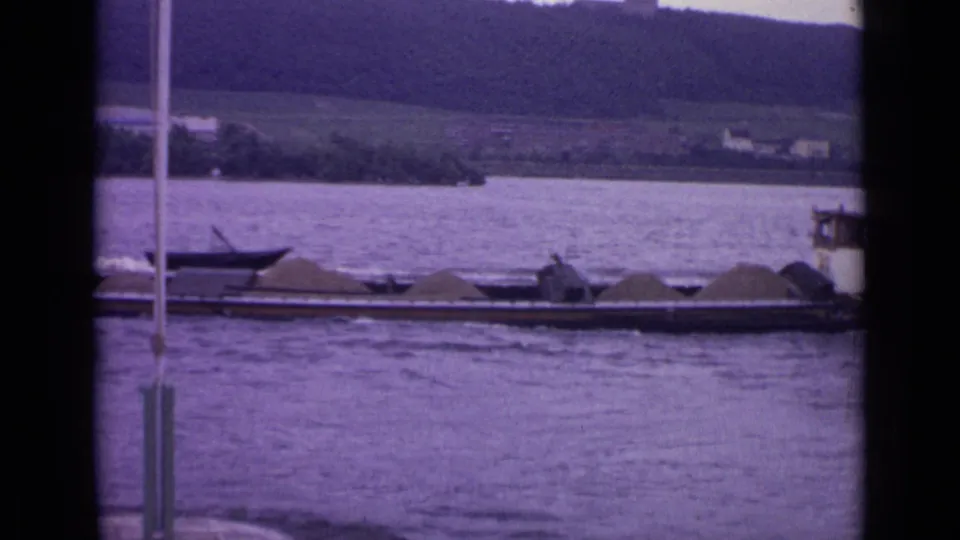 1962 Germany 8mm frame of a loaded river barge and small boats below a wooded hillside, with vertical window or railing elements in front.