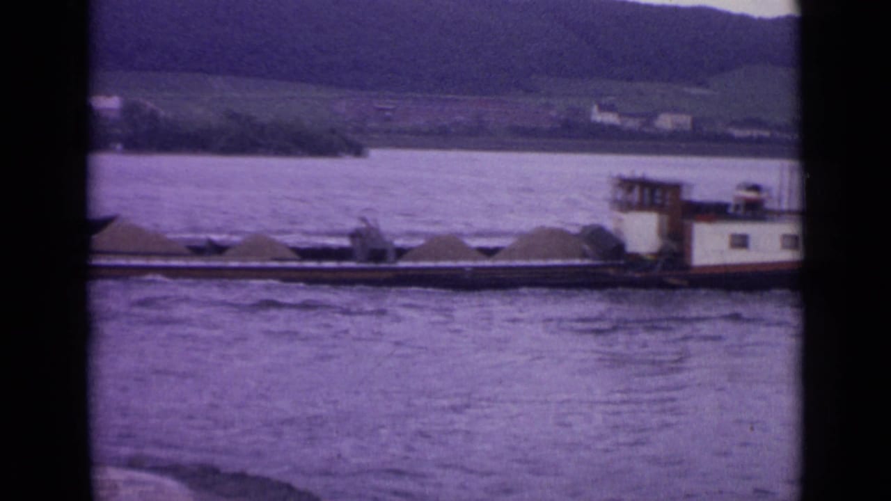 River barge carrying sand on a wide waterway with hills and buildings beyond.