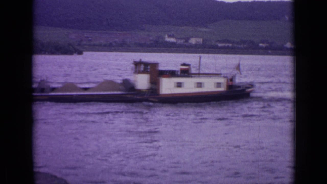 1962 Germany frame of a working barge crossing a broad river or lake, with low buildings and green hills visible across the water.