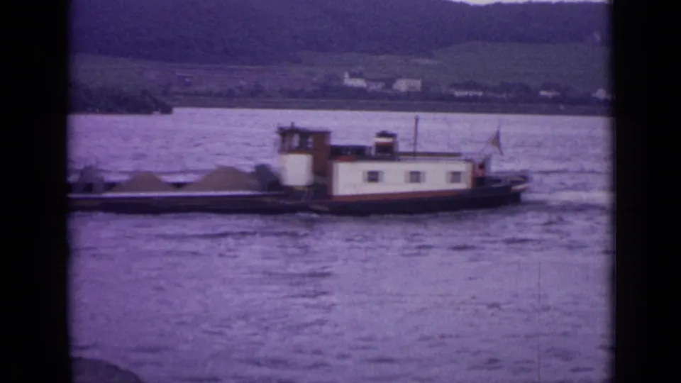 1962 Germany frame of a working barge crossing a broad river or lake, with low buildings and green hills visible across the water.