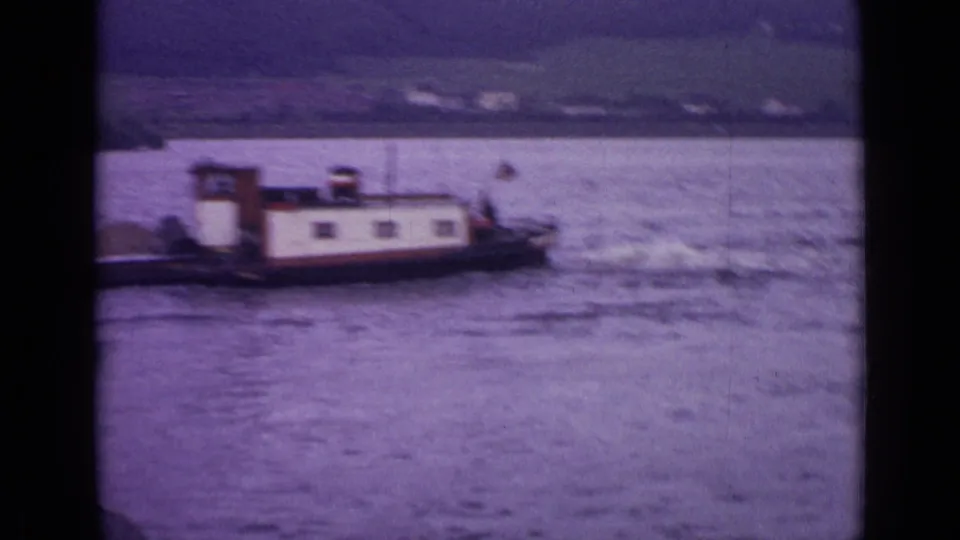 Soft 8mm frame showing the barge moving out of view across a broad German waterway, with a blurred hillside shoreline behind it.