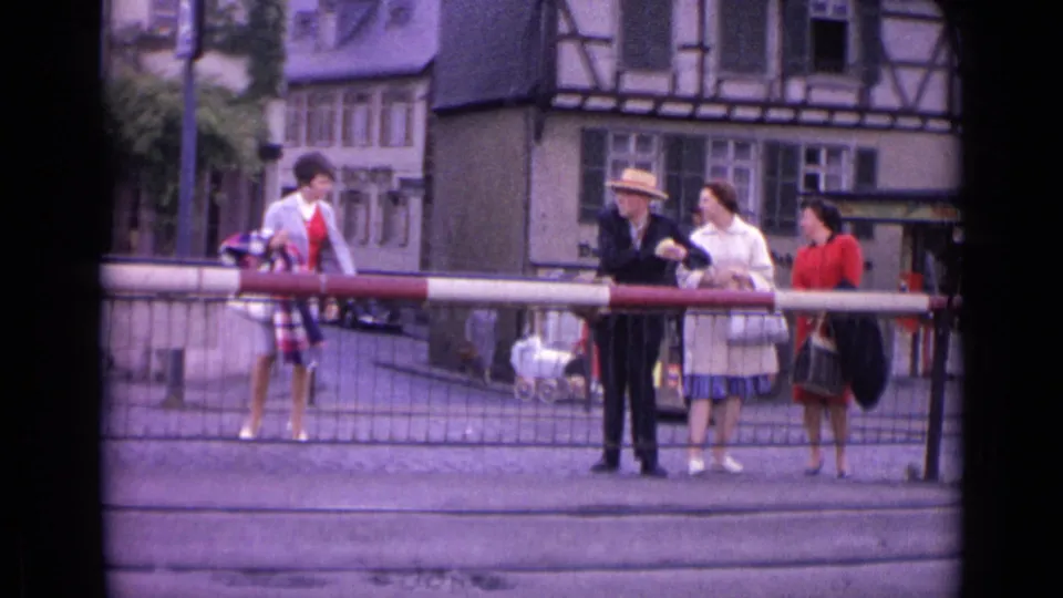 1962 Germany street scene with adults in period clothing waiting by a barrier near half-timbered buildings, including a man in a straw hat and a pram.