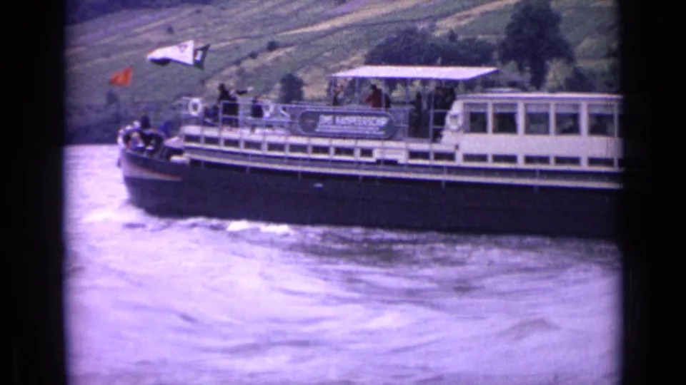 1962 German river scene with a passenger boat, visible deck passengers, flags, and terraced hills in the background.