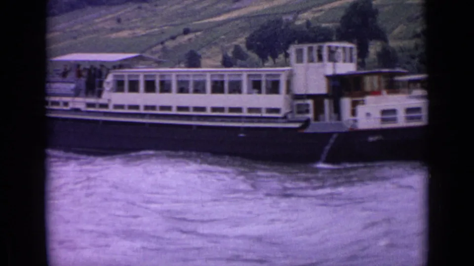 German 1962 8mm frame showing the side of a river passenger boat with passengers, cabin windows, and moving water.