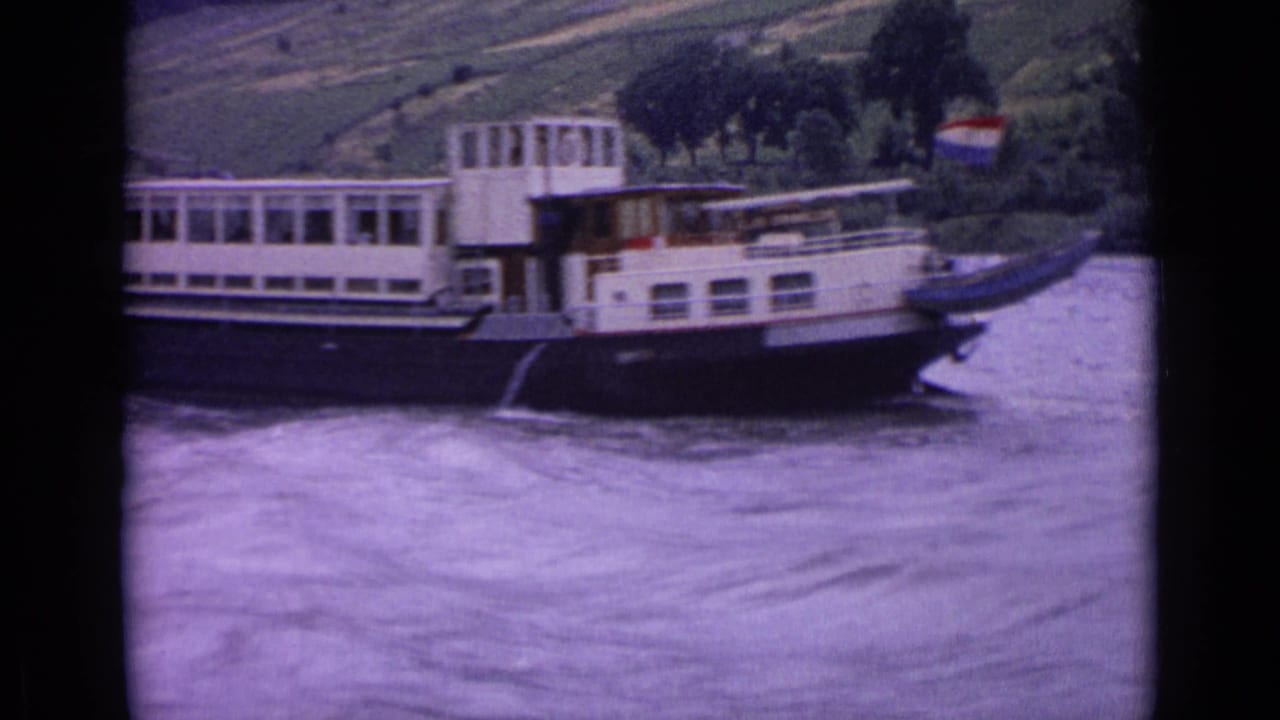 1962 Germany 8mm frame showing a white and dark-hulled riverboat passing on a broad river below a green hillside.