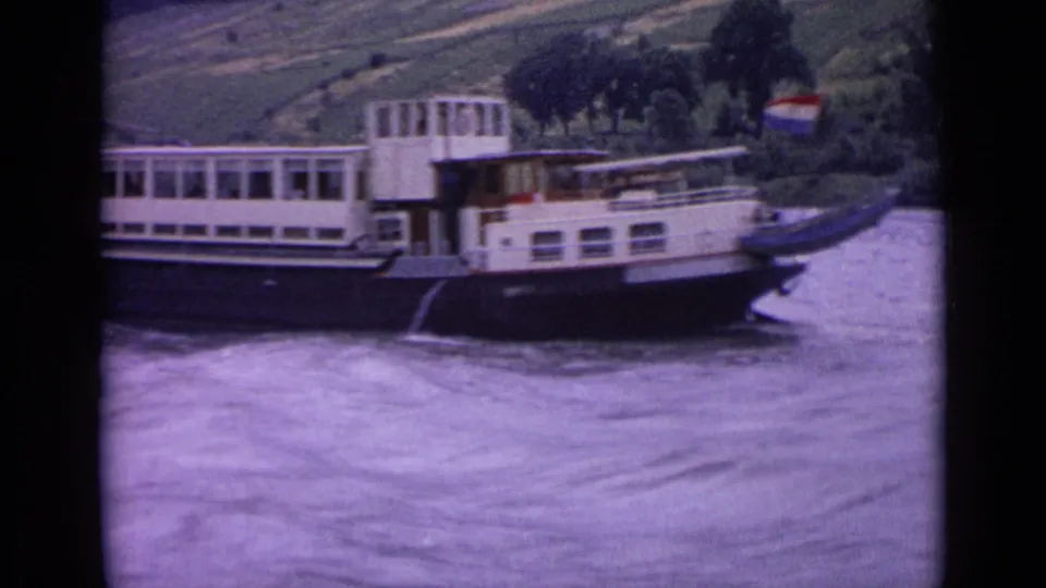 1962 Germany 8mm frame showing a white and dark-hulled riverboat passing on a broad river below a green hillside.