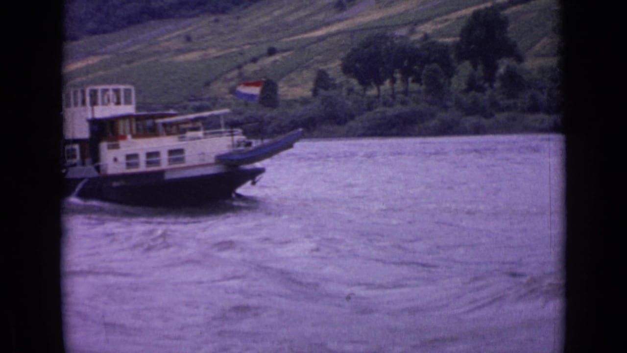 The riverboat continues downstream in 1962 Germany footage, with a small flag visible and the hillside receding behind it.