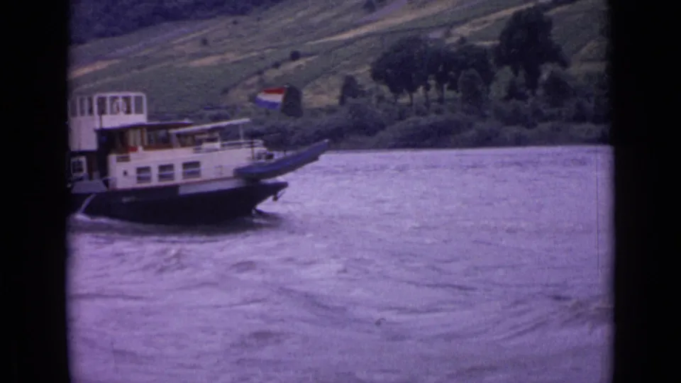 The riverboat continues downstream in 1962 Germany footage, with a small flag visible and the hillside receding behind it.