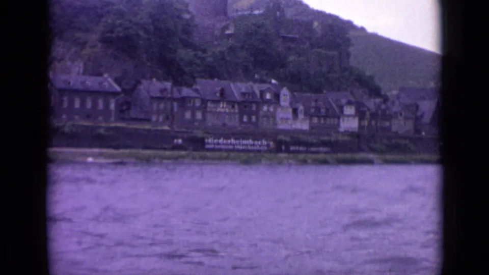 A German riverside town appears across the water, with clustered buildings at the foot of a tree-covered hill in 8mm footage.