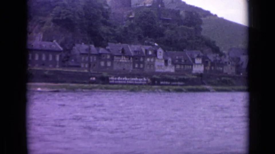 The final frame holds on the riverside town in Germany, showing water in the foreground and hillside trees above the buildings.