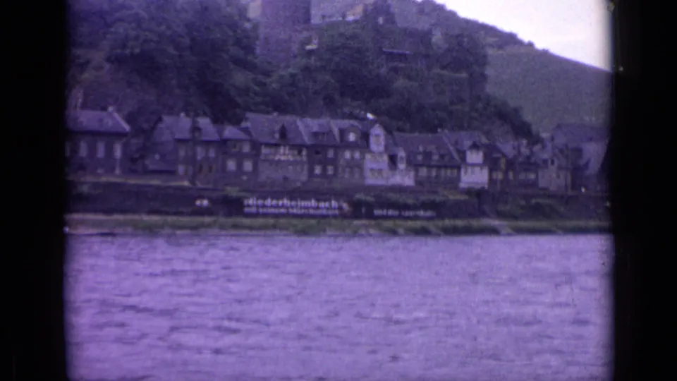 1962 8mm view of a German riverside town with traditional buildings, water in the foreground, and a castle hillside above.