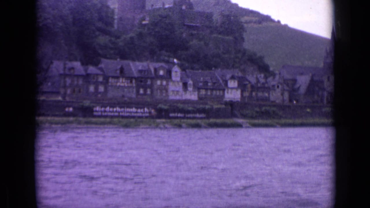 1962 German home-movie frame showing river water, closely packed town buildings, and a castle or ruins on the wooded hill.