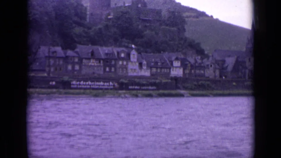 1962 German home-movie frame showing river water, closely packed town buildings, and a castle or ruins on the wooded hill.