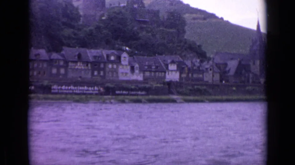 Vintage 8mm frame of a German riverside town, with half-timbered architecture, river foreground, hillside greenery, and a church steeple edge.