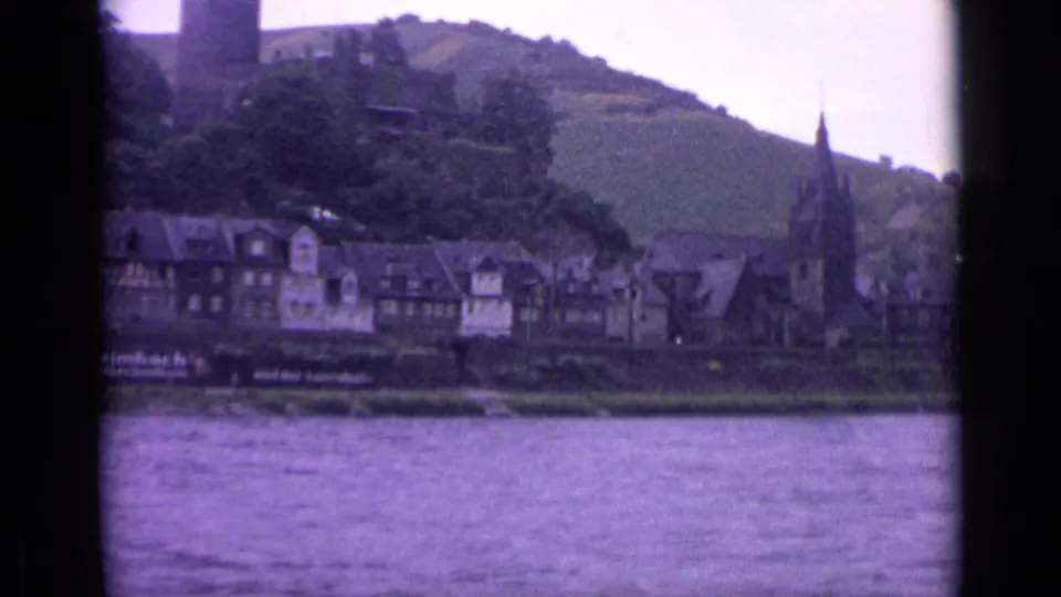 1962 Germany travel footage frame with river in front, a row of town buildings, a church steeple, and castle remains on the hill.