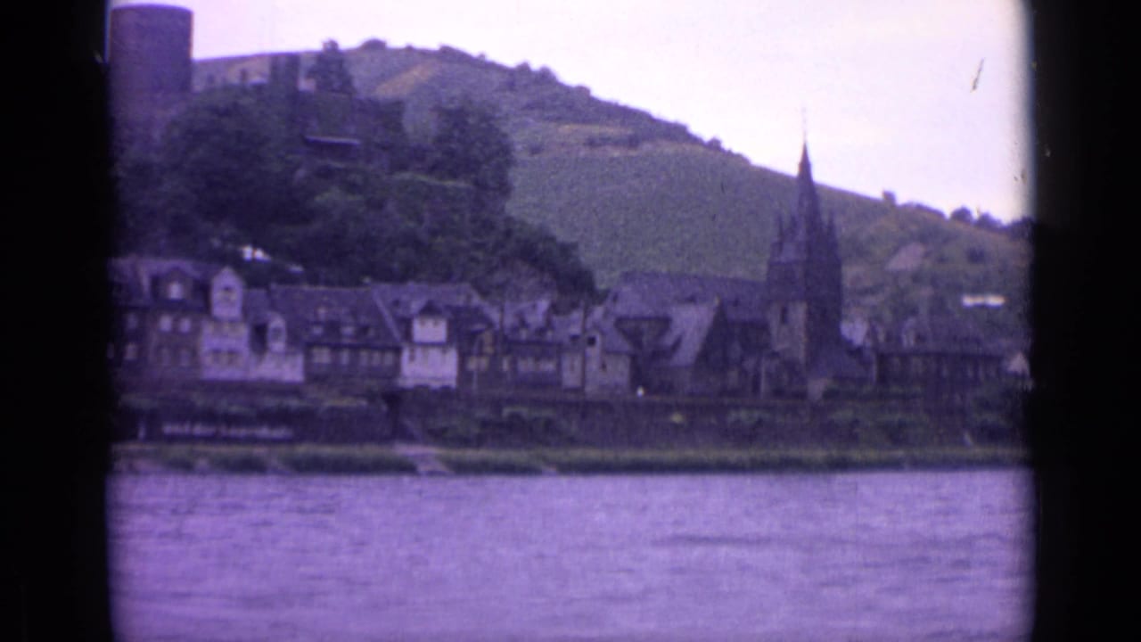 1962 8mm view of a German riverside town, with water in front, half-timbered buildings, a church steeple, and a green hillside behind.