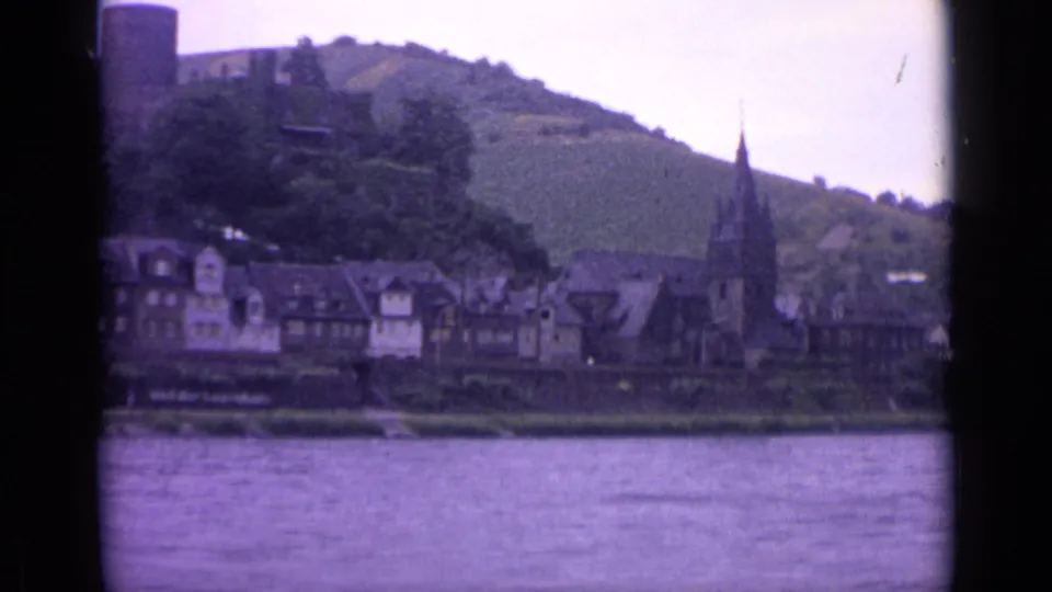 1962 8mm view of a German riverside town, with water in front, half-timbered buildings, a church steeple, and a green hillside behind.
