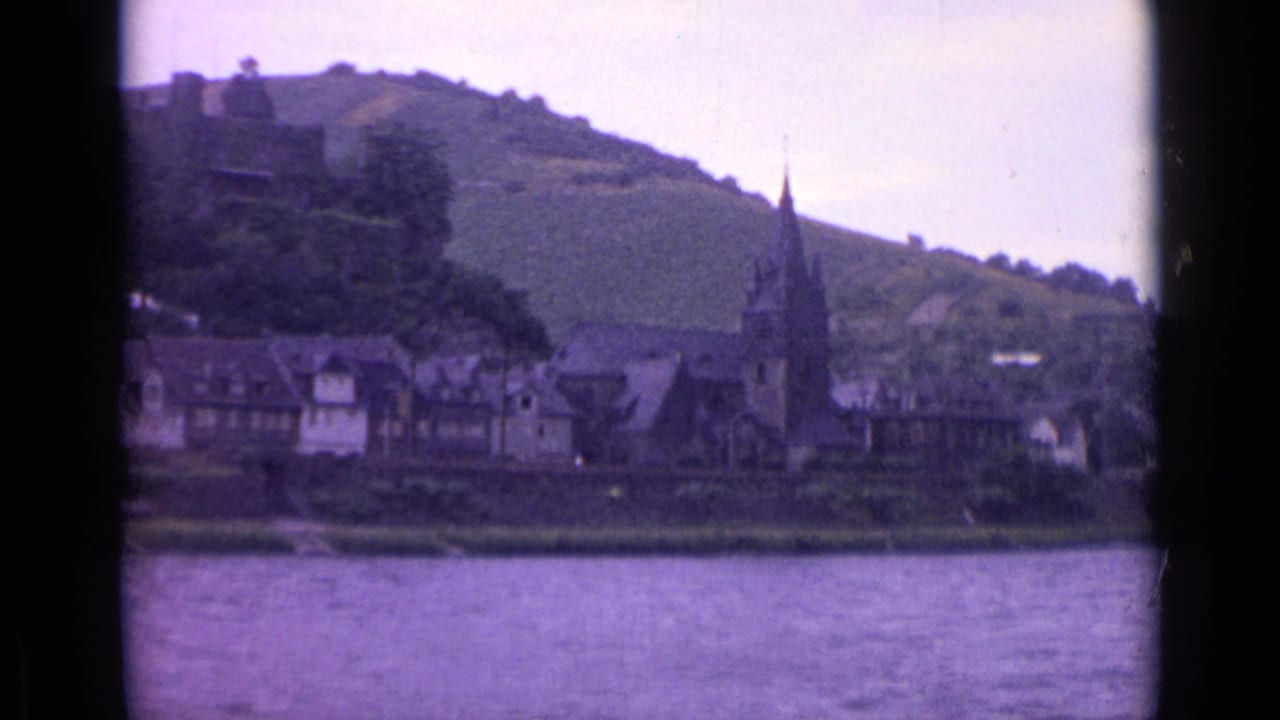 1962 German riverside view showing clustered buildings and a church steeple below a terraced green hill, filmed from across the water.