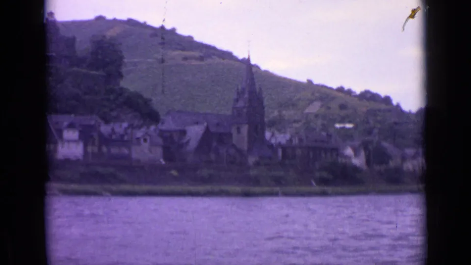 Soft 8mm frame from Germany in 1962 with a church steeple near center, riverside buildings, water foreground, and hillside rising behind.