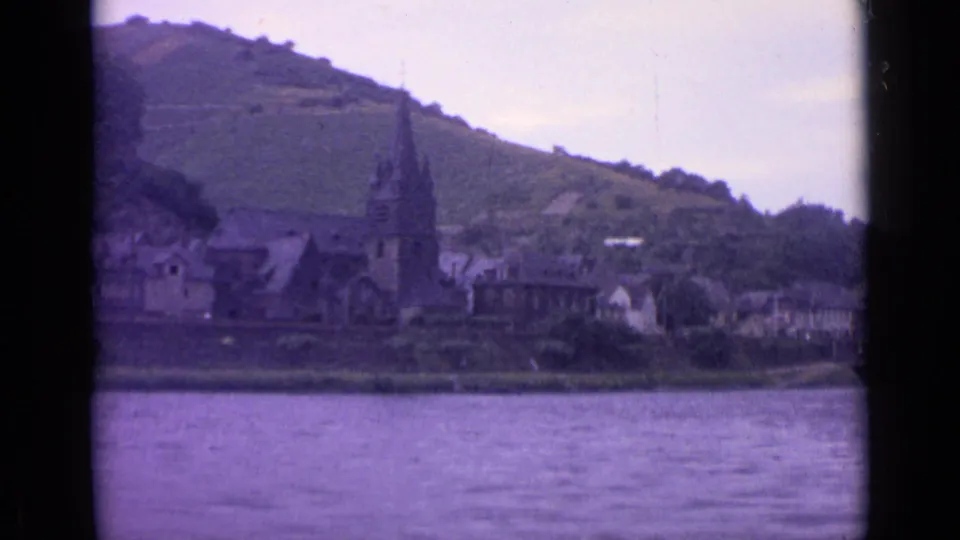1962 8mm frame of a German riverbank settlement, with church architecture, nearby houses, water in the foreground, and a cultivated hillside behind.