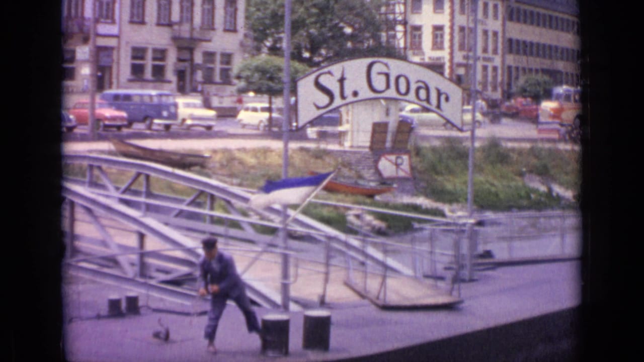 At the St. Goar pier in 1962 Germany, a man handles a rope near a metal gangway, with cars, buildings, and a small boat behind him.