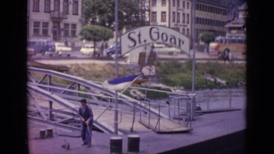 A man stands with a rope at the St. Goar pier in 1962, with metal gangway rails, a small boat, cars, trees, and town buildings behind him.
