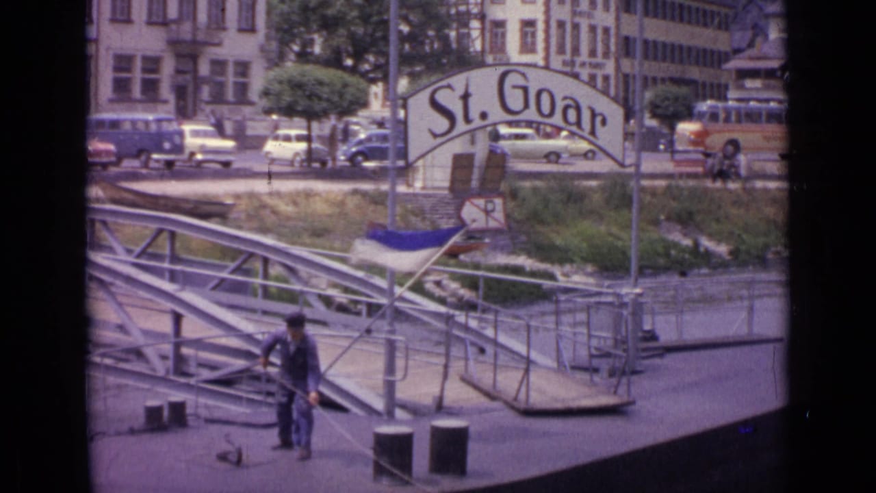 1962 Germany 8mm frame showing a man on a metal riverside dock pulling a rope below a large St. Goar sign, with cars and buildings behind.