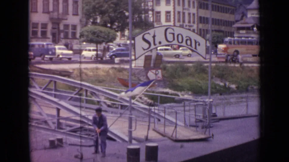 1962 Germany frame of a man holding rope on a riverside floating dock, with the St. Goar sign, cars, bus, and buildings in the background.