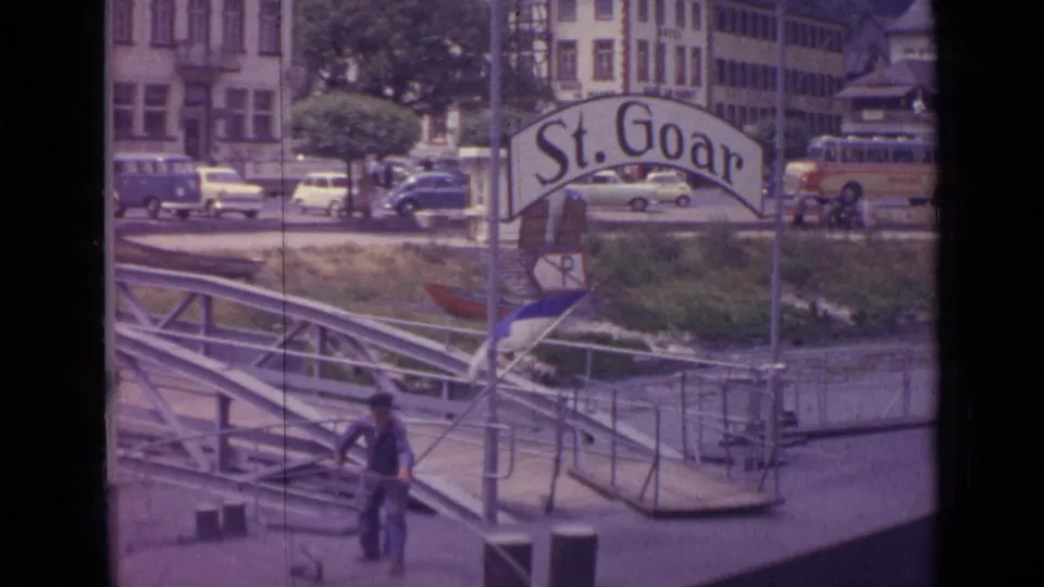 1962 Germany 8mm view of a man bending over a rope on the floating dock below the St. Goar sign, with cars and riverbank buildings visible.