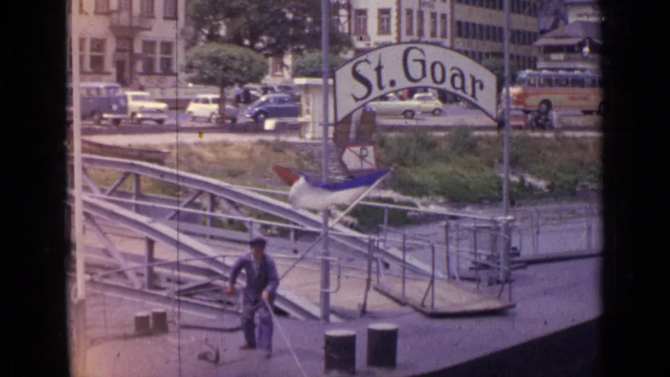 1962 Germany home-movie frame showing a man pulling rope on a metal floating dock beneath the St. Goar sign, with parked cars and buildings beyond.
