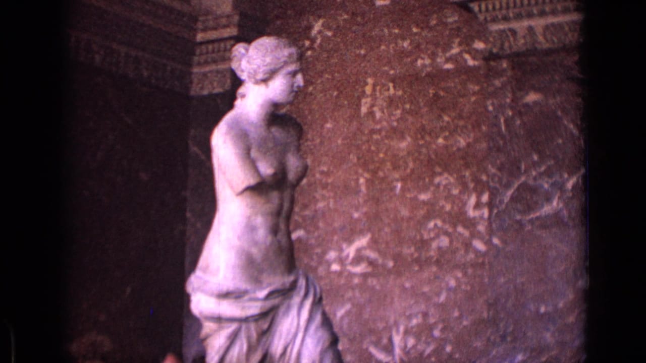 1962 8mm frame of the Venus de Milo statue in a grand museum hall, with stone wall panels and dark architectural recesses.