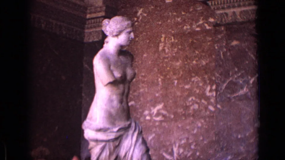 1962 8mm frame of the Venus de Milo statue in a grand museum hall, with stone wall panels and dark architectural recesses.