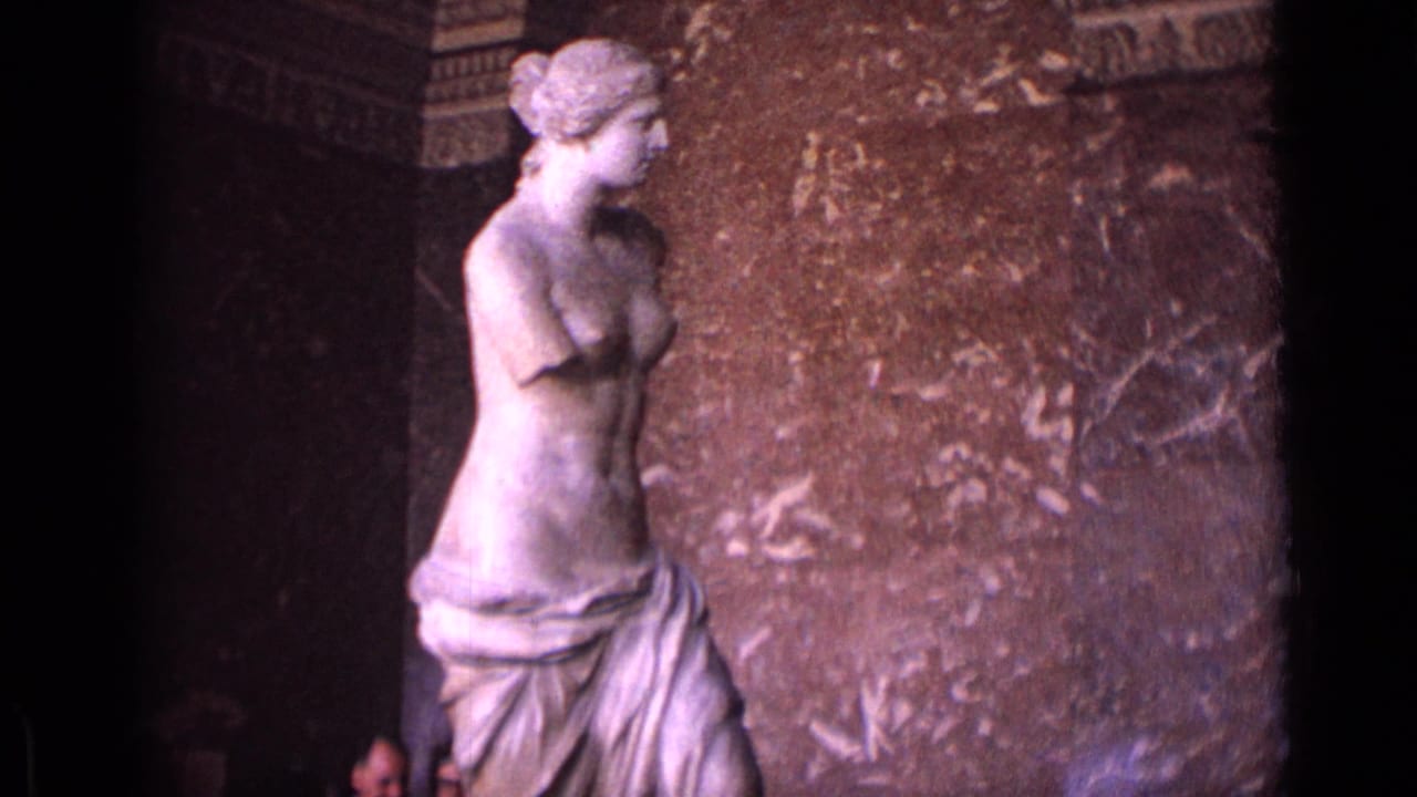The Venus de Milo remains centered in soft 8mm color, with mottled marble or stone wall surfaces behind it.