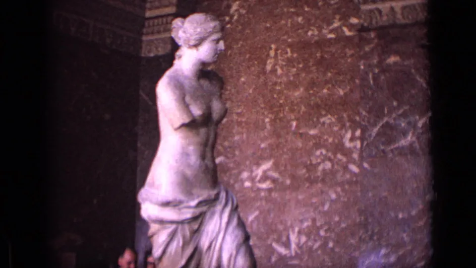 The Venus de Milo remains centered in soft 8mm color, with mottled marble or stone wall surfaces behind it.